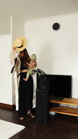 A girl in a straw hat and sunglasses holding a child poses with a suitcase in a room. Vertical video. The family is preparing for a trip. Air travel.