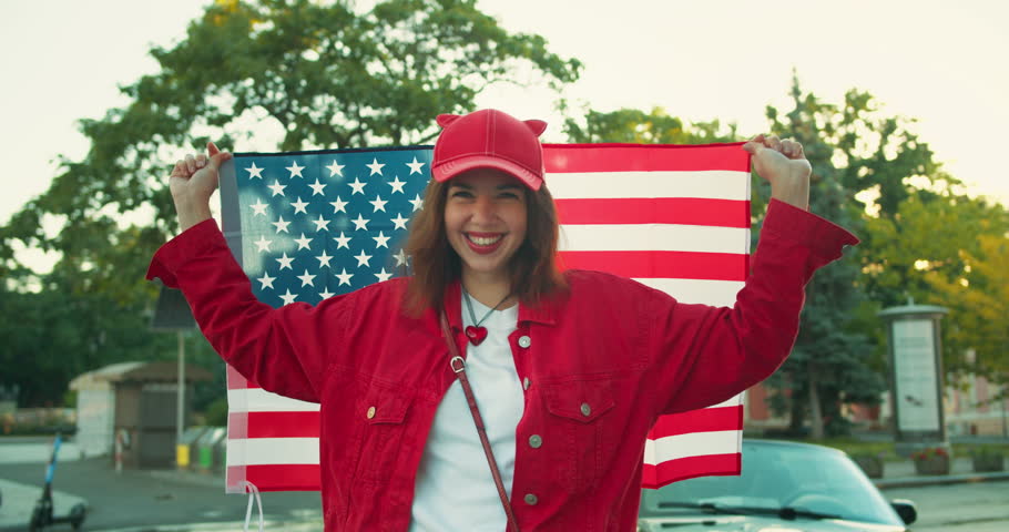 Close-up portrait of a happy cheerful girl in red clothes who holds a large US flag behind her back and is wrapped in it on the street