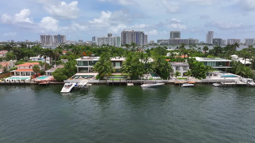 Waterfront homes in Miami Beach with private docks and boats. Luxurious properties surrounded by palm trees. Skyline of high-rise buildings in the background. Aerial.