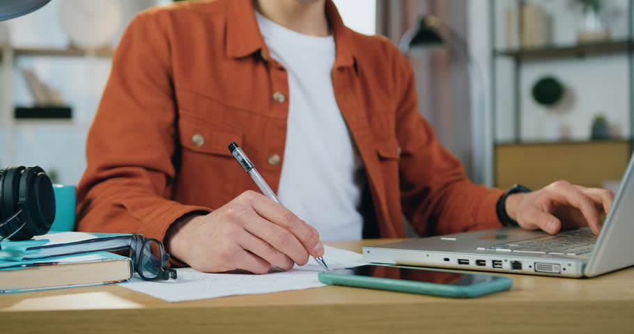 Close up. Unrecognizable diligent man student sitting near the computer and making needed notes into copybook while studying alone at home