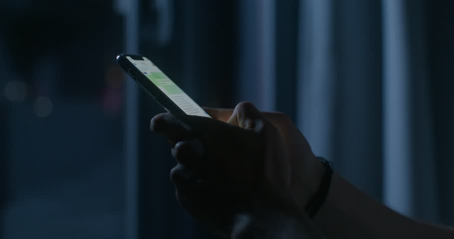 A man uses a mobile phone while sitting on the bed at night. Close-up of his hands, an unrecognizable person. Dark room, home interior