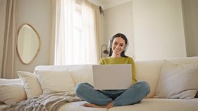 Zooming in on Caucasian woman using digital devices. Happy woman positively smiling while holding laptop on her legs. Sitting on sofa in living room at home. Wearing headphones while watching video. - Powered by Shutterstock - Get 15% off with code: PIKWIZARD15