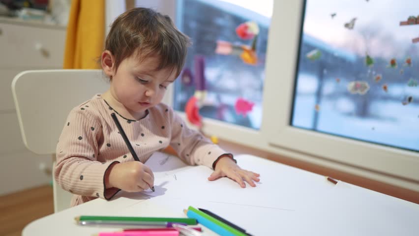 baby girl draws while sitting at table by the window at home. happy family kid concept. baby daughter learns to draw with pencils on a dream sheet of paper indoors. development of fine motor skills