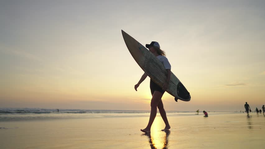 Beautiful girl surfer walking on picturesque ocean beach in sunset, slow motion. Active rest in summer vacation, activities on sea coastline, sport and recreation, traveling to oceanic shore, tourism