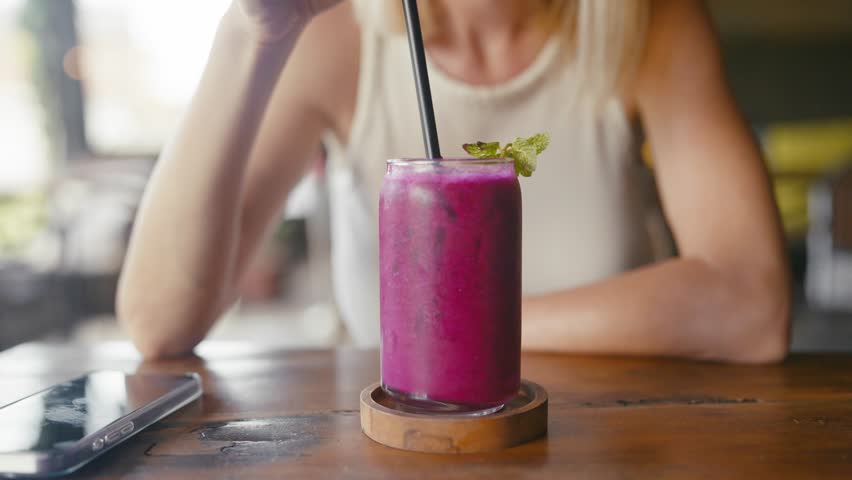 Young adult woman tourist enjoying exotic cocktails in Thai cafe in summer day. Closeup of cup with purple fruit smoothie on wooden table, lady stirring drink by plastic cocktail straw, relaxation