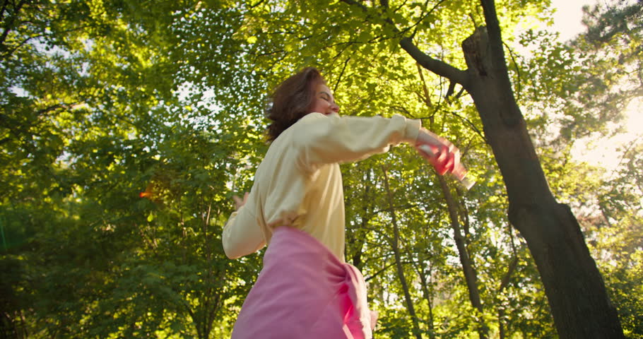 Happy brunette girl in a yellow hoodie with a pink shirt dances a cheerful dance and listens to music on wireless headphones in a green sunny park with sun rays