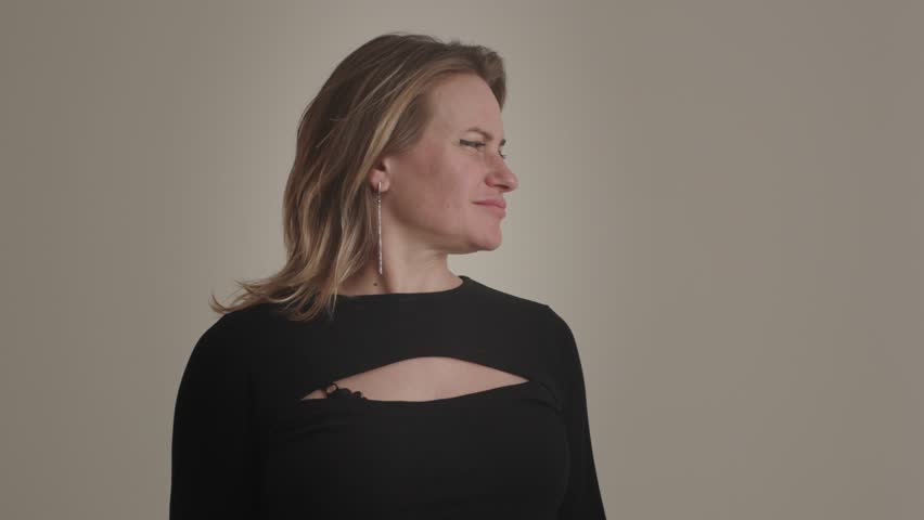 A charming young american or european blonde woman model poses in the studio for advertising or professional portfolio on the beige background. An elegant coquettish lady turns head, smiles, puts hand