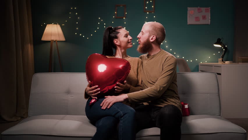 Young couple in love holding heart shaped balloon sitting on sofa in living room kissing while spending romantic evening together at home. Copy space
