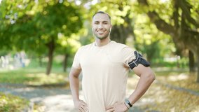 Portrait of a confident young sporty man smiling in park during an outdoor workout session. Man wearing casual athletic attire with armband and earphones, standing confidently with hands on hips - Powered by Shutterstock - Get 15% off with code: PIKWIZARD15