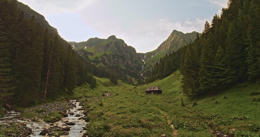 Establishing aerial view of a scenic mountain waterfall with an idyllic wooden cabin and a mountain river in the background. Concept of scenic landscape, nature retreat, and outdoor living