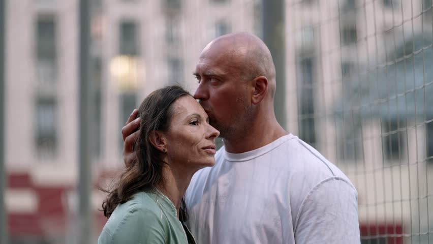 An adult couple in love hugging tenderly, standing at a street stadium in the city. A bald man with an athletic build, a woman with a soft smile, look happy and confident.