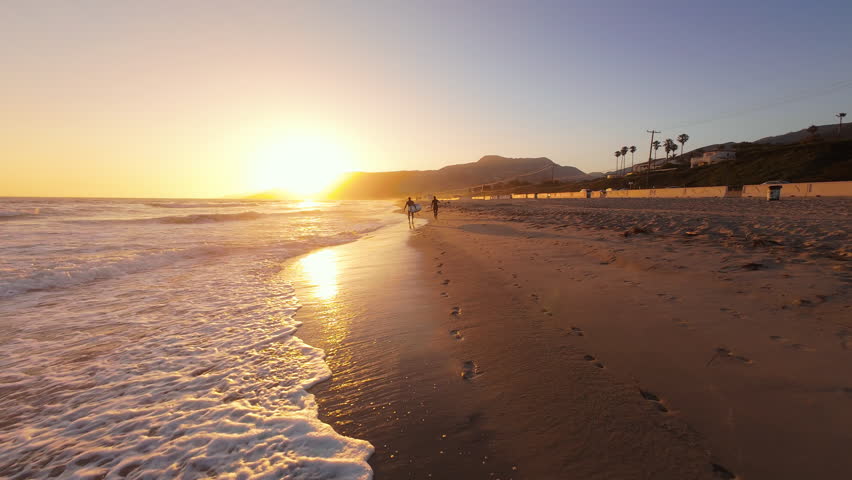 Captured by an FPV drone, a beautiful sunset paints a serene scene on a sandy Malibu beach, with waves, mountains, and strolling figures completing the idyllic evening landscape
