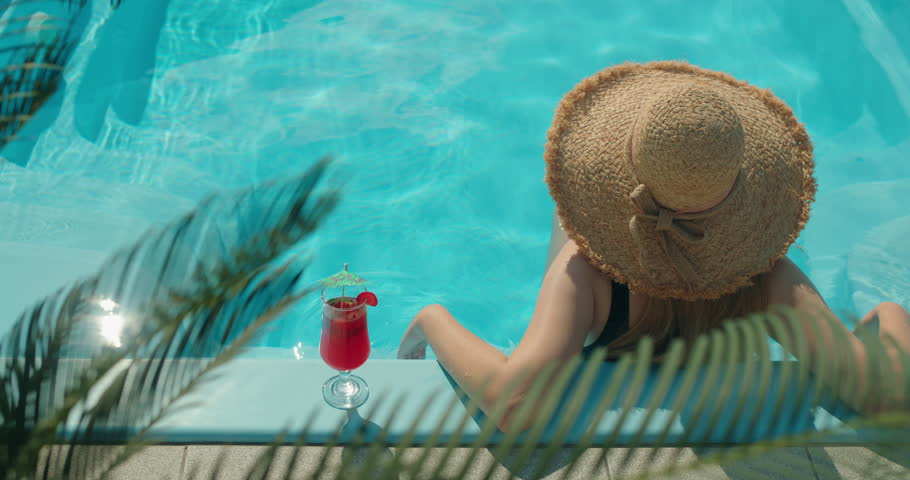 Teenage girl in a straw hat relaxing by the pool, palm leaves swaying in the foreground