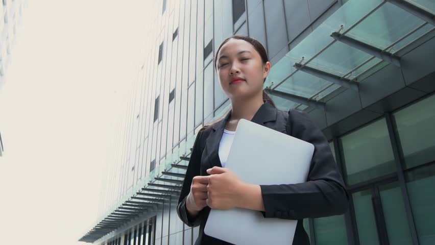 Asian businesswoman standing holding laptop and smiling at camera behind tall building