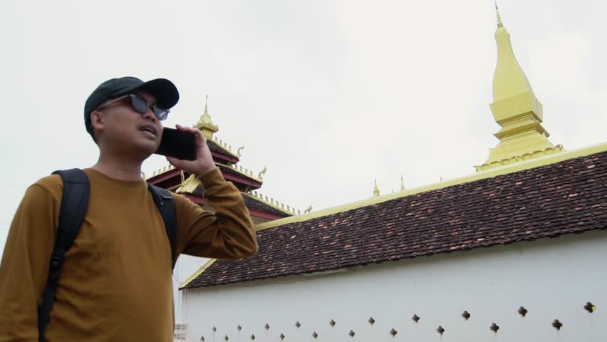 Asian male tourist using phone to talk with friend about traveling to see the art and culture of Lao PDR. Pha That Luang, the concept of cultural tourism in Laos.