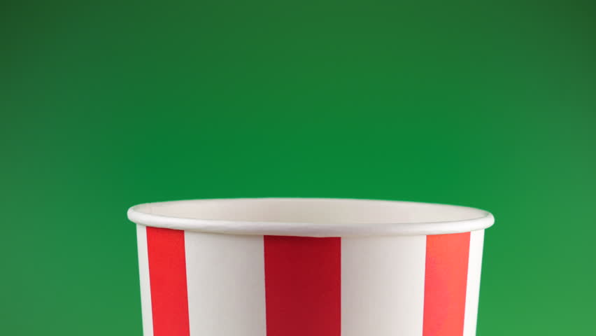 Popcorn grains falling into red and white striped bucket isolated on green background. Movie snack food, fast food cafe sweet snack. Slow motion. Chromakey or chroma key background.
