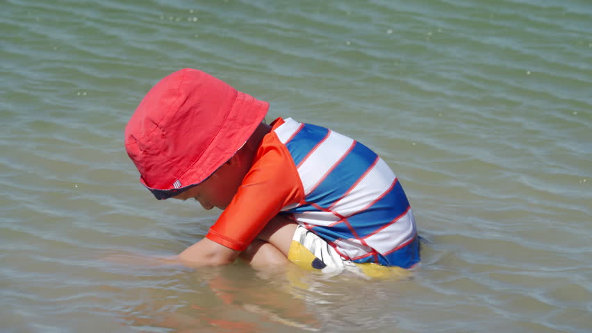 Small child wearing sun protection shirt playing on the lake shore on a summer day.