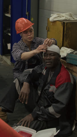 Vertical full shot of young Hispanic workwoman putting bandage on coworkers head, administering first aid to male Black factory worker after labor accident in plant