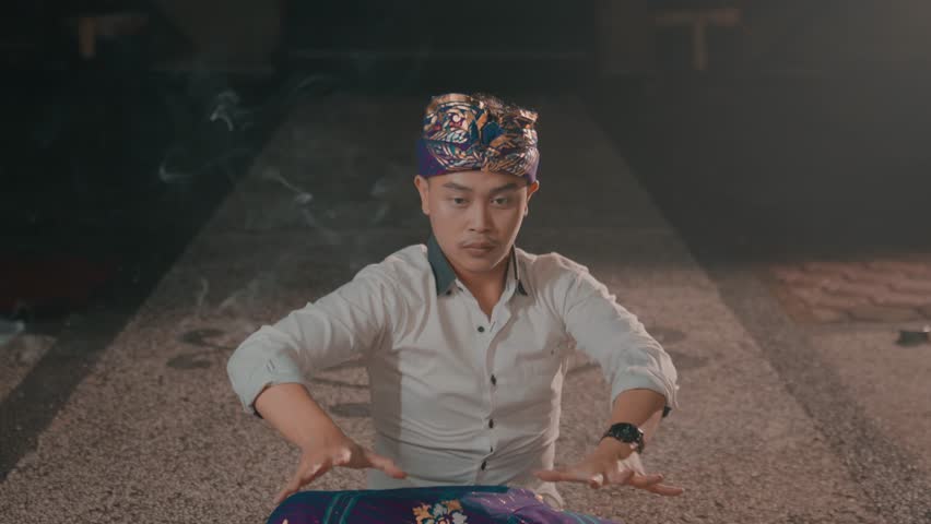 A person wearing traditional attire and a headband is performing a ritual or dance with hands raised in a dimly lit setting.
