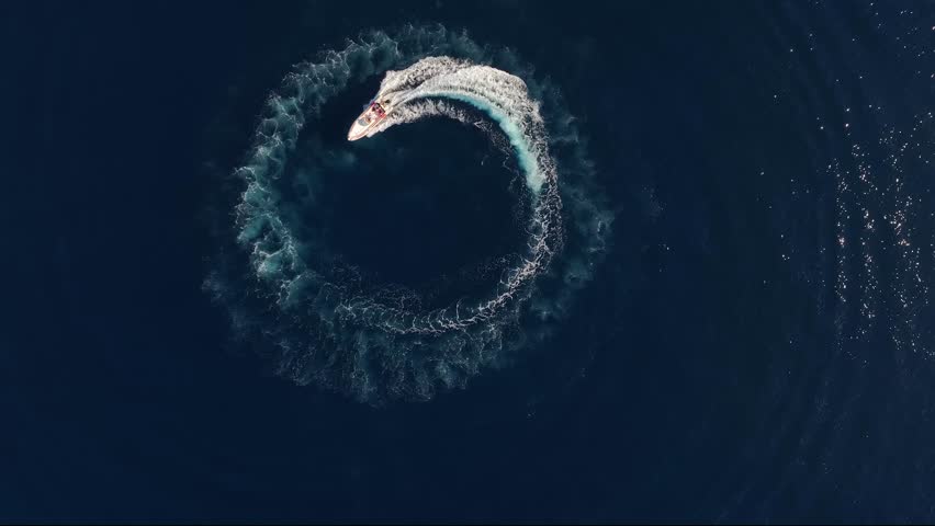 Aerial top view of a motor RIB boat forming a circle of waves and bubbles with its engine over the blue sea