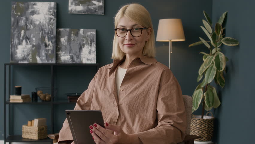 Medium portrait of confident Caucasian female psychologist using digital tablet while sitting on chair at her modern bright office, posing for camera