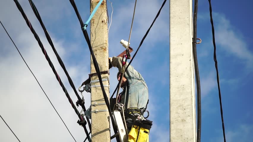 Skilled electrician in helmet fixes wires standing on ladder near high pole against blue sky on summer day backside view. Electrical service and mounting on the pole. Slow motion
