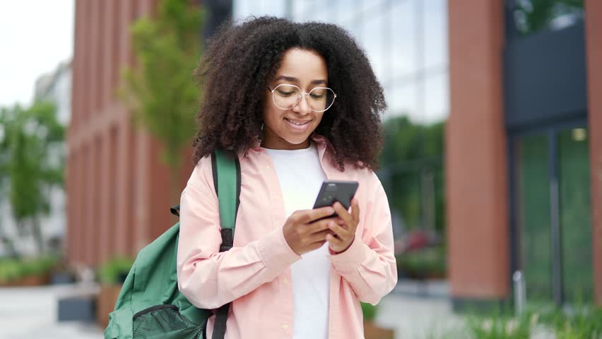 Smiling african american female student using phone standing on campus space near university building. A happy black woman with backpack reads writes messages, browses social networks or chats online