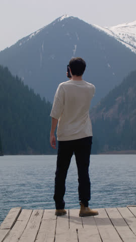 Vertical: man on a wooden pier on a mountain lake. A guy travels alone among the mountain peaks, an adventure. Male uses a smartphone, takes selfies. Blue water in the lake, untouched nature