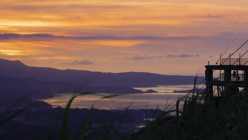 A vibrant sunset displays a mix of yellow and orange hues over a coastal radar station. The silhouette of the radar equipment contrasts with the colorful sky and distant mountains, Taiwan.