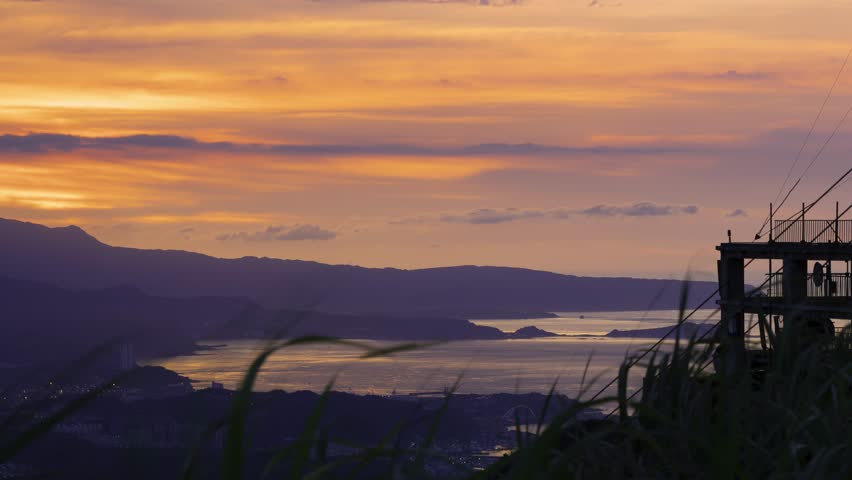 A vibrant sunset displays a mix of yellow and orange hues over a coastal radar station. The silhouette of the radar equipment contrasts with the colorful sky and distant mountains, Taiwan.