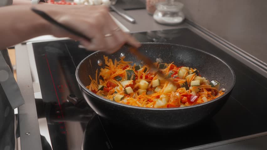 A woman mixes grated carrots, zucchini, onion, and bell pepper with a spatula in a non-stick frying pan on an induction stovetop. The colorful vegetables are combined evenly for a healthy dish.