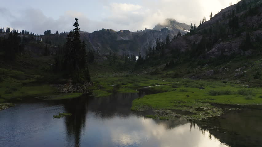 Rocky PNW alpine landscape reflects in shimmering meadow lake with lush green forest and grass, aerial dolly
