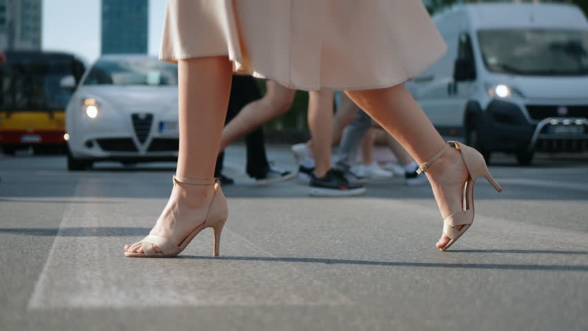 Following female feet steps crossing busy street. Slim woman wearing dress and high heels goes on pedestrian crossing in city. Low angle people crowd legs walking. No visible brand names and logos