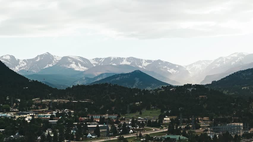 Drone rises above Estes Park Colorado as sunlight breaks between clouds across snow covered Rocky Mountains