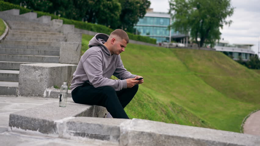 in the park young guy in a gray sweatshirt sits on a stone staircase chatting on the phone cloudy cool weather