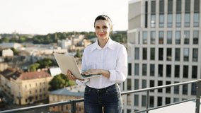 Smiling woman holding laptop and giving thumbs up on rooftop with cityscape background. Woman wearing casual white shirt and jeans, representing modern remote work lifestyle. - Powered by Shutterstock - Get 15% off with code: PIKWIZARD15
