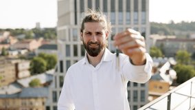 Smiling man in white shirt holding keys while standing outdoors with modern city buildings in background. Concept of real estate, new home, and achievement. - Powered by Shutterstock - Get 15% off with code: PIKWIZARD15