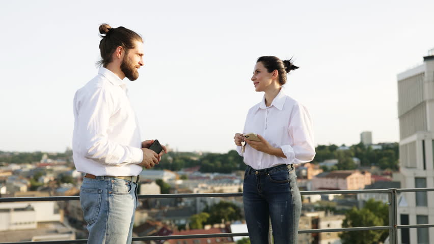 Real estate agent handing keys to new homeowner on rooftop with cityscape background. Man and woman in casual attire smiling, holding electronic devices, and celebrating new property transaction. - Powered by Shutterstock - Get 15% off with code: PIKWIZARD15