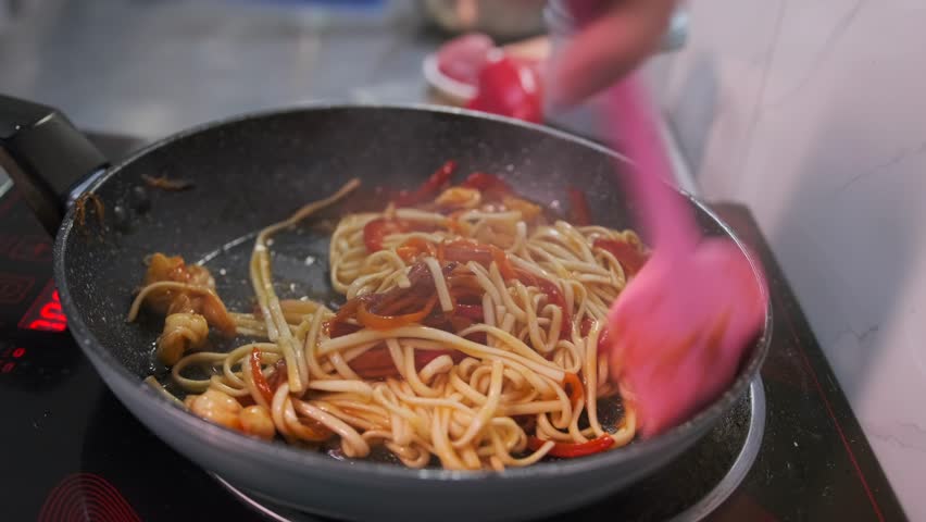 A hand stirs with a spatula noodles with shrimp and tomatoes wok udon fried in a frying pan on an electric stove, close-up.
