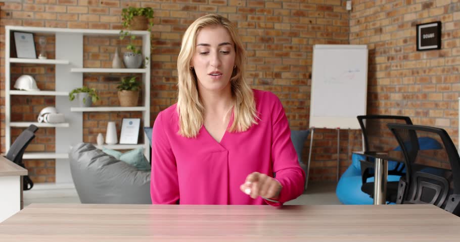 Close up of people talking to the camera, businesswoman in pink blouse discussing work at office desk, gesturing. the person is fully visible, sitting, half body, pointing at things on a table