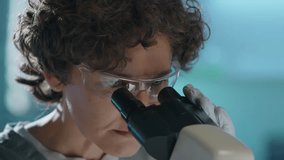 Close-up tilt shot of Caucasian female researcher in protective glasses placing specimen on glass slide under objective lens to study under microscope, then looking into ocular, while working in lab - Powered by Shutterstock - Get 15% off with code: PIKWIZARD15