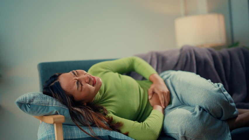 Young woman in severe abdominal pain, clutching stomach and transitioning from sitting to lying on a couch. Expressions of discomfort and agony