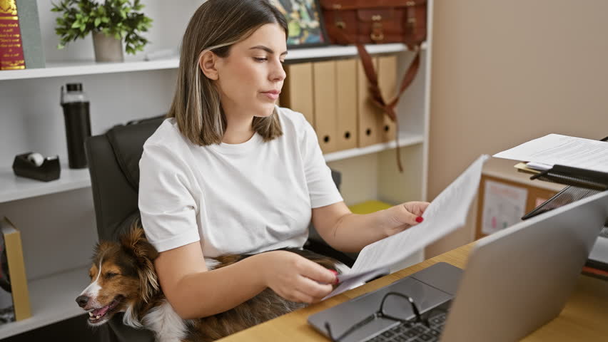 Hispanic woman works in office with glasses, laptop, papers, dog, multitasking, organized, white shirt, and plant.