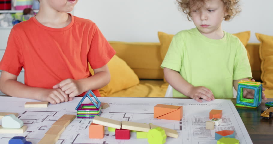 Boy shows room on plan to little brother holding magnet constructor details at table. Toddler kid refuses to build house with friend in playroom