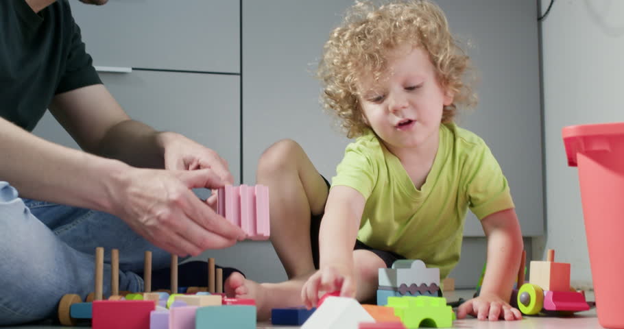 Smart little boy places constructor blocks onto tower with father sitting on floor. Happy child learns to build pyramid with loving parent at home