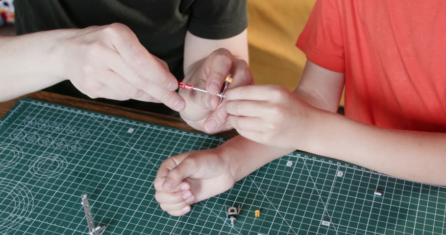 Father screws while little son holds metal details of constructor set at table. Man and toddler boy make machine model of steel parts in playroom