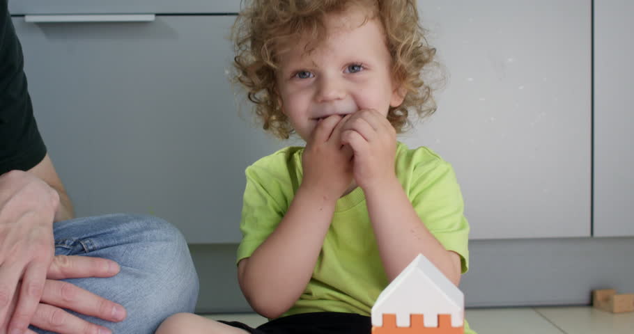 Baby child finishes tower of wooden blocks while father applauds on floor closeup. Happy littler son builds house of constructor set with parent