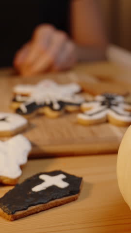 Vertical video. A Ghost-Shaped Cookie in Black Icing, Homemade Treats for Halloween. A Woman Offers It to Me, with Many Other Cookies Shaped Like Pumpkins and Skeletons in Coffins on the Table.