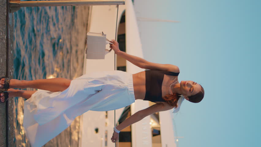 Fashionable model standing sea quay in elegant trendy outfit vertical video. Beautiful confident woman posing in front luxury yacht moored at port. Chic girl relaxing at summer. Carefree vacation