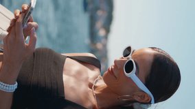 Charming girl looking smartphone at sunny seaside vertical video closeup. Happy elegant woman laughing reading cellphone message on sea coast. Stylish lady in sunglasses watching social media at shore - Powered by Shutterstock - Get 15% off with code: PIKWIZARD15